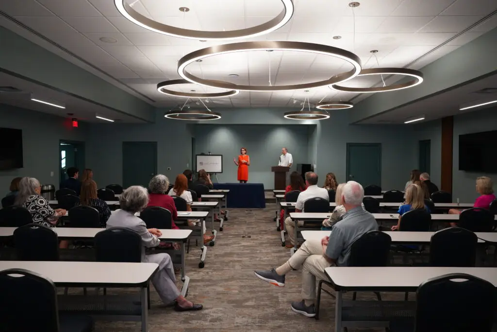 An indoor presentation or ceremony taking place in a large conference room with teal walls, long rows of classroom-style tables, and distinctive large, circular ring pendant lights hanging from the ceiling. At the front of the room, a woman in a bright orange dress speaks next to a presentation screen displaying the East Alabama Community Foundation logo, while a man stands nearby at a wooden podium. An audience is seated at the tables, listening to the presentation.
